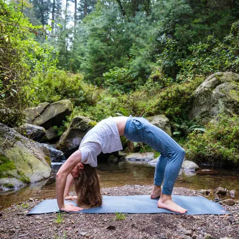 A male yogi performing a half wheel pose in front of a mountain stream, with a lush, green forest in the background
