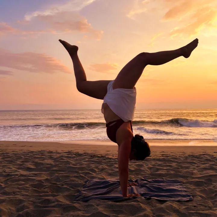 A female yogi performing a hanstand with legs spread out on a beach during sunset